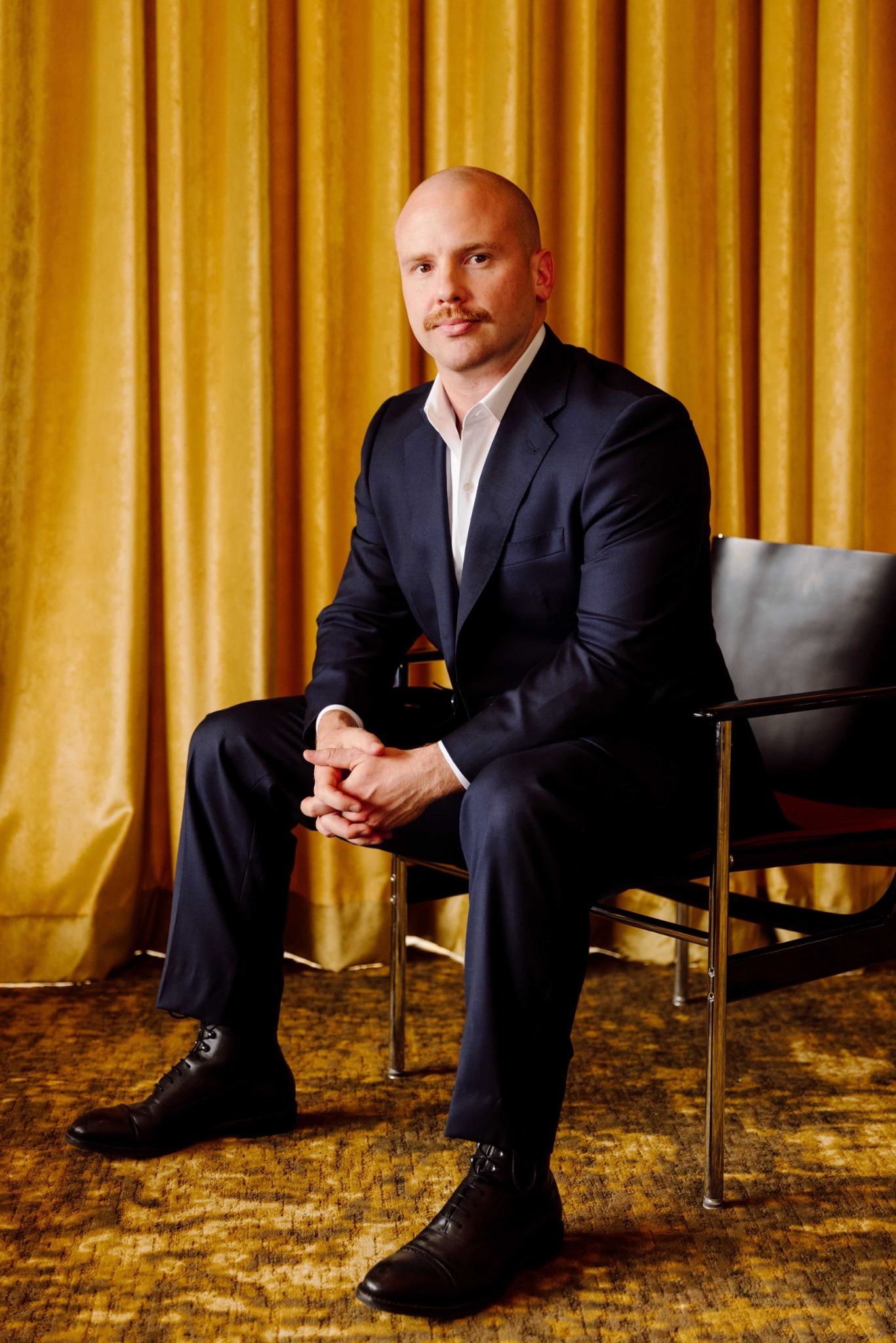 Kyle Tut seated in a navy suit in front of gold curtains